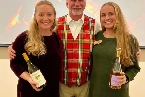 3 people stand together in festive holiday attire, holding wine bottles and glasses, with a fireplace projected onto a screen behind them 
